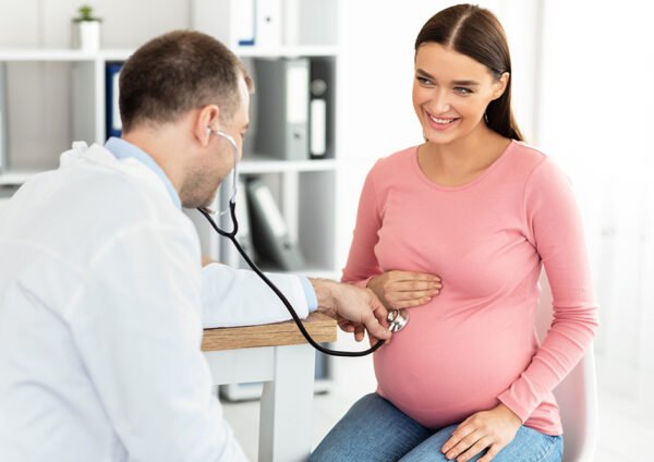Gynecology Consultation. Smiling pregnant woman visiting her obstetrician doctor in maternity clinic, male gynecologist examining her belly with stethoscope, doing medical check-up